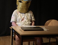 Eine Person sitzt an einem Tisch auf der Bühne und trägt eine Tüte auf dem Kopf. 