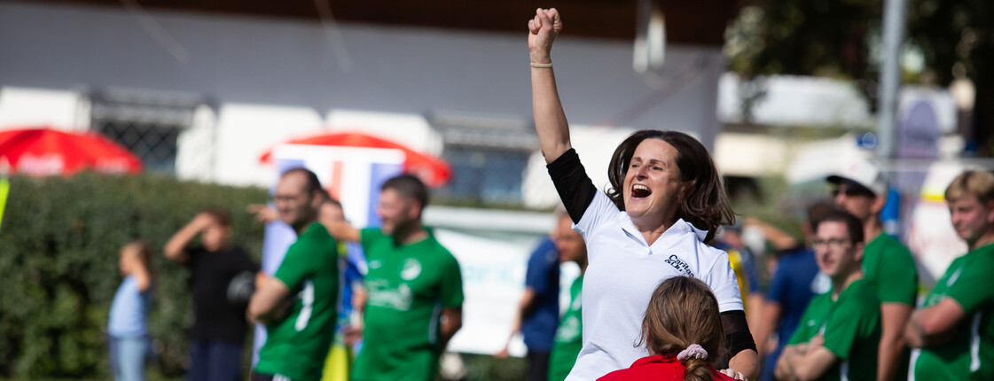 Eine Frau in einem weißen T-Shirt jubelt bei einem Fußballspiel am Fußballplatzrand. Es sind einige Fußballspieler in grünen Dressen zu sehen.