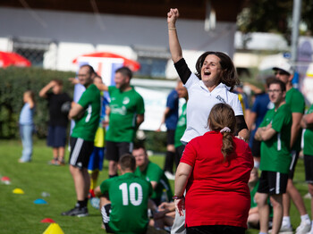 Eine Frau in einem weißen T-Shirt jubelt bei einem Fußballspiel am Fußballplatzrand. Es sind einige Fußballspieler in grünen Dressen zu sehen.