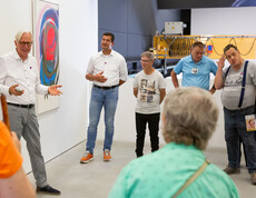 Ein Mann spricht vor einer Gruppe von Besuchern in einem Ausstellungsraum mit zeitgenössischer Kunst.