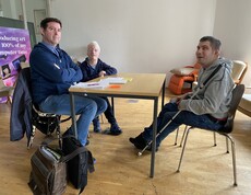 Zwei Männer und eine Frau sitzen am Tisch und schauen in die Kamera. 