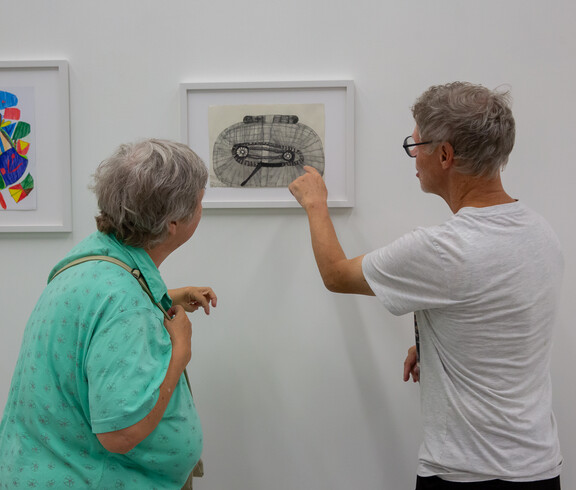 Ein Mann erklärt einer Frau in einem Ausstellungsraum ein von ihm gezeichnetes Bild.