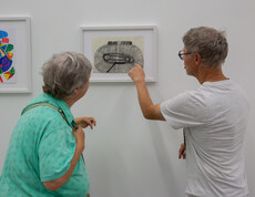 Ein Mann erklärt einer Frau in einem Ausstellungsraum ein von ihm gezeichnetes Bild.