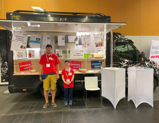 Ein Mann und eine Frau stehen vor einem Food Truck der mit Infomaterial dekoriert ist. 