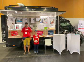 Ein Mann und eine Frau stehen vor einem Food Truck der mit Infomaterial dekoriert ist. 
