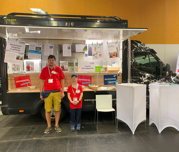 Ein Mann und eine Frau stehen vor einem Food Truck der mit Infomaterial dekoriert ist. 