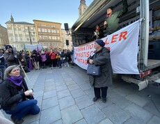 Eine Frau steht vor einem Wagen und spricht zu einem Publikum auf einem öffentlichen Platz.