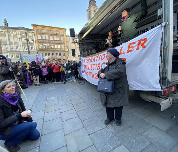 Eine Frau steht vor einem Wagen und spricht zu einem Publikum auf einem öffentlichen Platz.