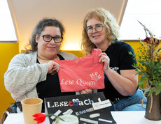Zwei Frauen halten ein T-Shirt, auf dem Lese Queen drauf steht, in die Kamera. Vor ihnen auf dem Tisch steht ein Schild, auf dem Lesecoach steht.
