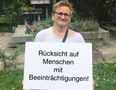 Eine Frau hält ein Schild auf dem steht "Rücksicht auf Menschen mit Beeinträchtigungen"