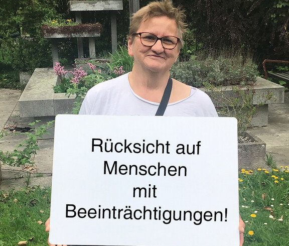 Eine Frau hält ein Schild auf dem steht "Rücksicht auf Menschen mit Beeinträchtigungen"