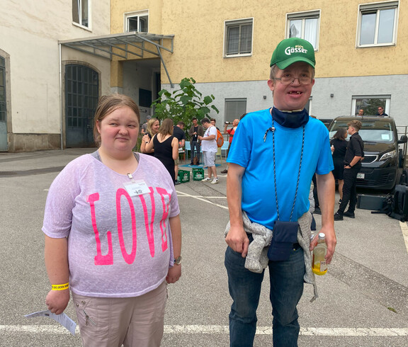 Eine Frau in einem rosa Shirt und ein Mann in einem blauem Shirt stehen vor einem Gebäude. 