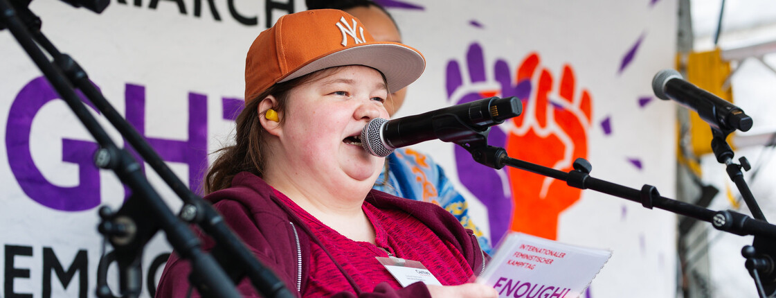 Eine junge Frau mit Kappe hält auf einer Bühne zum Weltfrauentag eine Rede