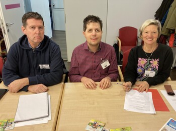 Zwei Männer und eine Frau sitzen an Tischen bei einer Pressekonferenz