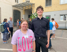 Eine Frau in rosa Shirt und ein Mann in schwarzem Hem stehen vor einem Gebäude. 