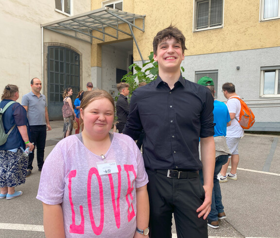 Eine Frau in rosa Shirt und ein Mann in schwarzem Hem stehen vor einem Gebäude. 