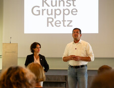 Eine Frau und ein Mann stehen vor einer Leinwand, auf der Kunst Gruppe Retz steht.