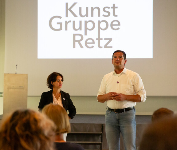 Eine Frau und ein Mann stehen vor einer Leinwand, auf der Kunst Gruppe Retz steht.