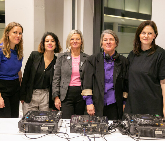 Fünf Frauen stehen hinter einem DJ Pult und schauen in die Kamera. 
