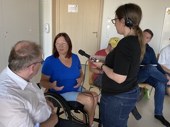 Ein Frau sitzt im Rollstuhl und spricht mit einem Mann während eine andere Frau ein Mikrofon hält.