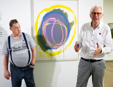 Ein Mann erklärt in einem Ausstellungsraum mit zeitgenössischer Malerei ein Kunstwerk. Im Hintergrund ist der Künstler zu sehen.