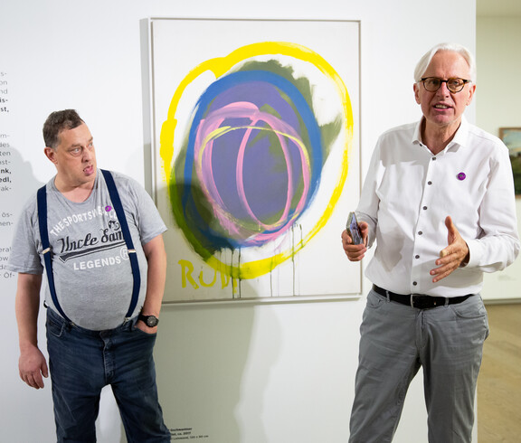 Ein Mann erklärt in einem Ausstellungsraum mit zeitgenössischer Malerei ein Kunstwerk. Im Hintergrund ist der Künstler zu sehen.