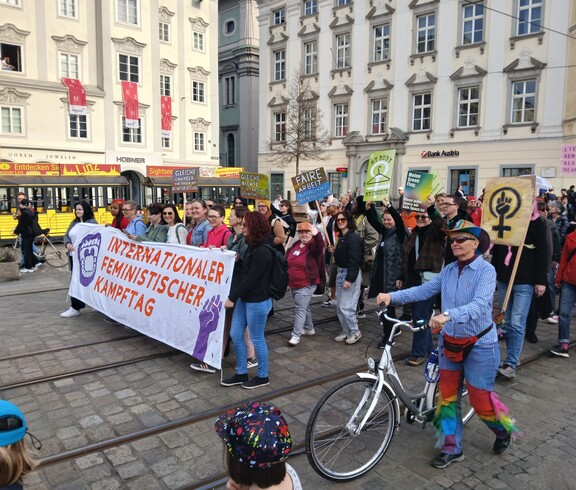 Demonstrationszug beim Feministischen Kampftag 2026 mit einem großen Banner im Vordergrund. Im Hintergrund Häuser am Linzer Hauptplatz.