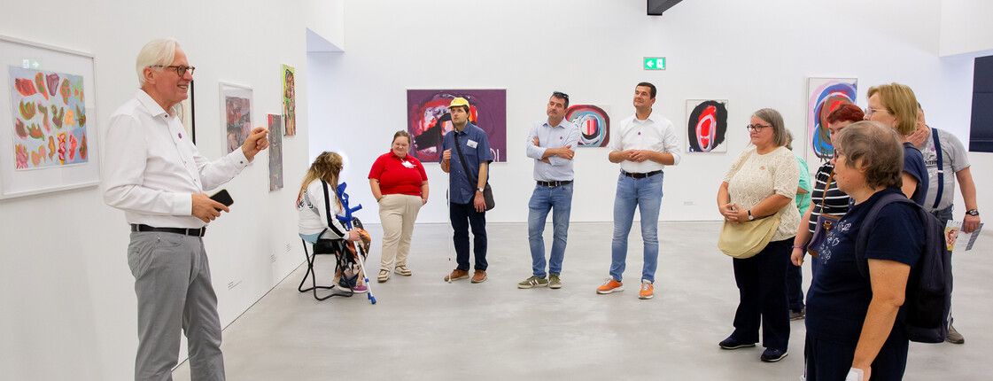 Ein Mann spricht vor einer Gruppe von Besuchern in einem Ausstellungsraum mit zeitgenössischer Kunst.