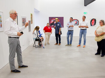 Ein Mann spricht vor einer Gruppe von Besuchern in einem Ausstellungsraum mit zeitgenössischer Kunst.