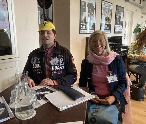 Ein Mann mit einer Blindenkappe und eine Frau sitzen an einem Tisch. 