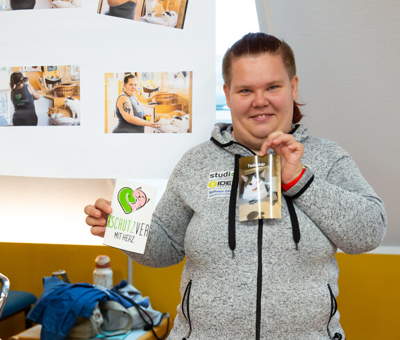 Eine junge Frau hält ein Foto einer Katze und ein Fotos mit "Tierschutzverein mit Herz"  in Händen. Vor ihr auf dem Tisch liegen einige Katzenfotos, hinter sind auf einem Plakat mehrere Fotos zu sehen, auf dem sie mit Katzen abgebildet ist.