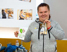 Eine junge Frau hält ein Foto einer Katze und ein Fotos mit "Tierschutzverein mit Herz"  in Händen. Vor ihr auf dem Tisch liegen einige Katzenfotos, hinter sind auf einem Plakat mehrere Fotos zu sehen, auf dem sie mit Katzen abgebildet ist.