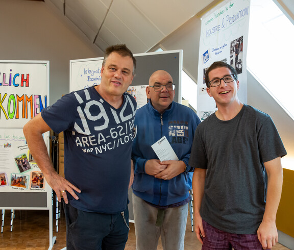 Drei Männer stehen nebeneinander vor zwei Pinwänden und einem Plakat. Auf der einen Pinwand steht "Herzlich Willkommen" drauf.