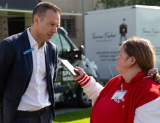 Ein Mann im Anzug wird von einer jungen Frau  mit einem Handy am Rande eines Fußballplatzes interviewt. Im Hintergrund stehen zwei kleine Trucks.