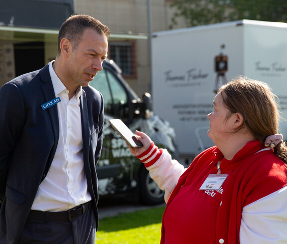 Ein Mann im Anzug wird von einer jungen Frau  mit einem Handy am Rande eines Fußballplatzes interviewt. Im Hintergrund stehen zwei kleine Trucks.