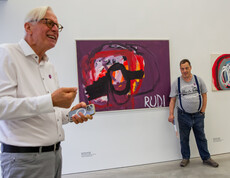 Ein Mann erklärt in einem Ausstellungsraum mit zeitgenössischer Malerei ein Kunstwerk. Im Hintergrund ist der Künstler zu sehen.