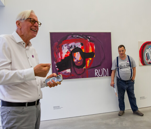 Ein Mann erklärt in einem Ausstellungsraum mit zeitgenössischer Malerei ein Kunstwerk. Im Hintergrund ist der Künstler zu sehen.