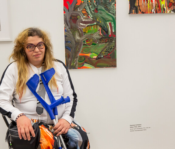 Eine Frau sitzt mit Krücken vor zwei ihrer Kunstwerke in einem Ausstellungsraum für zeitgenössische Kunst.
