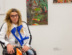 Eine Frau sitzt mit Krücken vor zwei ihrer Kunstwerke in einem Ausstellungsraum für zeitgenössische Kunst.