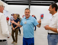 Ein Mann spricht zu zwei Männern in einem Ausstellungsraum für zeitgenössische Kunst. Im Hintergrund sind andere Ausstellungsbesucher zu sehen.