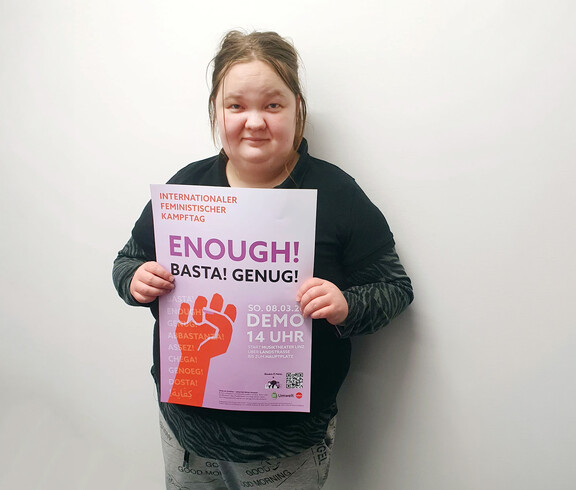 Eine Frau steht vor einer weißen Wand und hält ein Plakat in die Kamera.