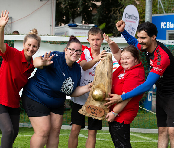 Drei Frauen und zwei Männer jubeln am Rande des Fußballplatzes von Blau Weiß Linz. Sie haben den CariCup-Pokal 2026 in den Händen.