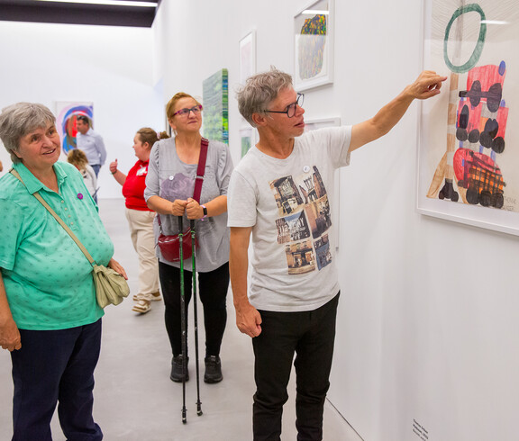 Ein Mann erklärt einer Frau in einem Ausstellungsraum ein von ihm gemaltes Bild. Im Hintergrund sind andere Ausstellungsbesucher zu sehen.