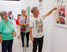 Ein Mann erklärt einer Frau in einem Ausstellungsraum ein von ihm gemaltes Bild. Im Hintergrund sind andere Ausstellungsbesucher zu sehen.