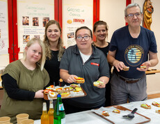 Vier Frauen und ein Mann präsentieren vor einem Brötchen-Buffet belegte Brötchen, die sie in Händen halten. Im Hintergrund sind handgeschriebene Plakate zu sehen.