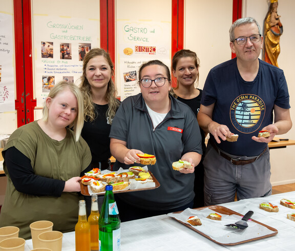 Vier Frauen und ein Mann präsentieren vor einem Brötchen-Buffet belegte Brötchen, die sie in Händen halten. Im Hintergrund sind handgeschriebene Plakate zu sehen.