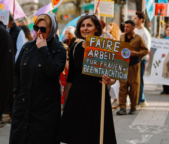 Zwei Frauen gehen bei der Demonstration am Weltfrauentag 2026 mit einem Schild mit. Auf dem Schild steht "Faire Arbeit für Frauen mit Beeinträchtigung"