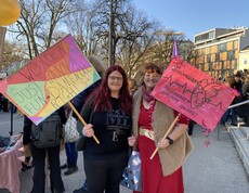 Zwei Frauen halten ein Plakat in der Hand und blicken in die Kamera.