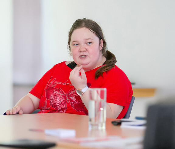 Eine Frau im rotem T-Shirt sitzt an einem Tisch und spricht.