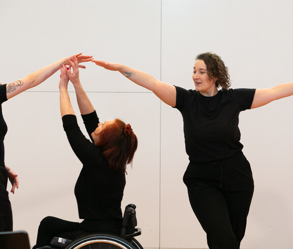 Drei Frauen tanzen vor einer weißen Wand. Eine davon ist im Rollstuhl. 